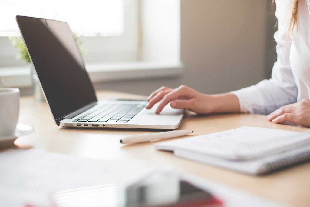 A businesswoman typing on a laptop at a bright indoor workspace with notebooks and a pen.